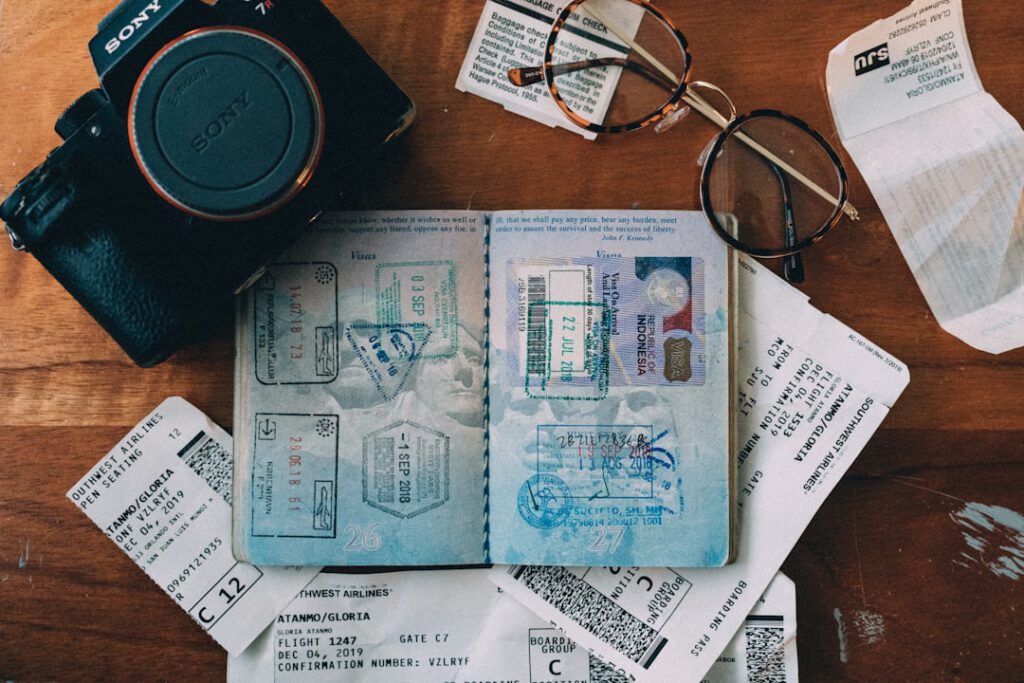 Open passport with visa stamps, boarding passes, camera, and glasses laid out on a wooden table — travel planning flat lay. Photo by Henry Thong.