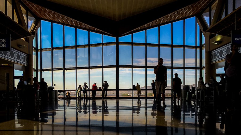 Silhouettes of travelers standing in a large sunlit airport terminal window, evoking the feeling of waiting during a flight delay.