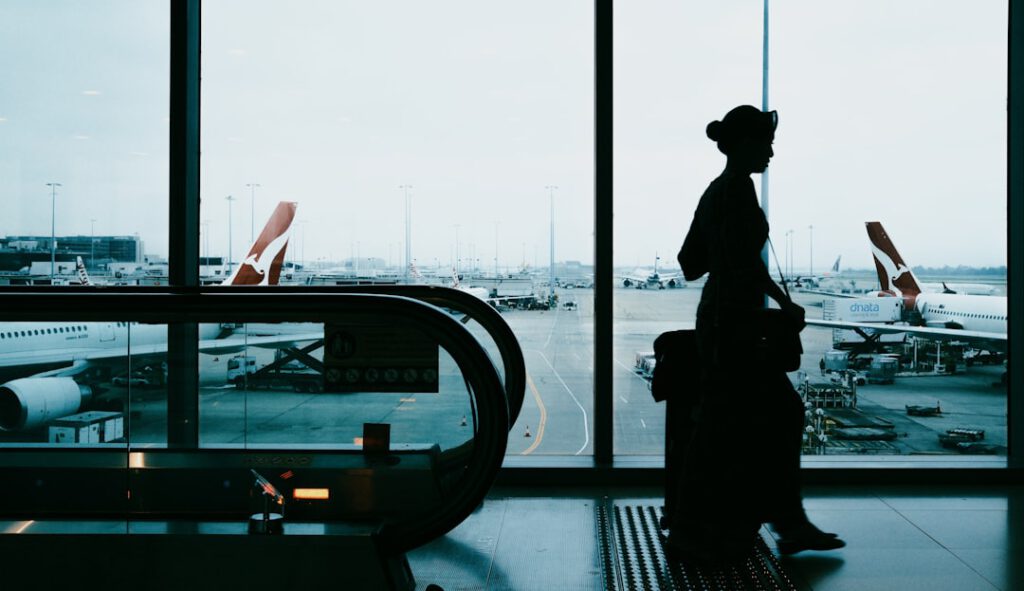 Silhouette of a traveler with suitcase standing at airport terminal window with planes on the tarmac — photo by Unsplash